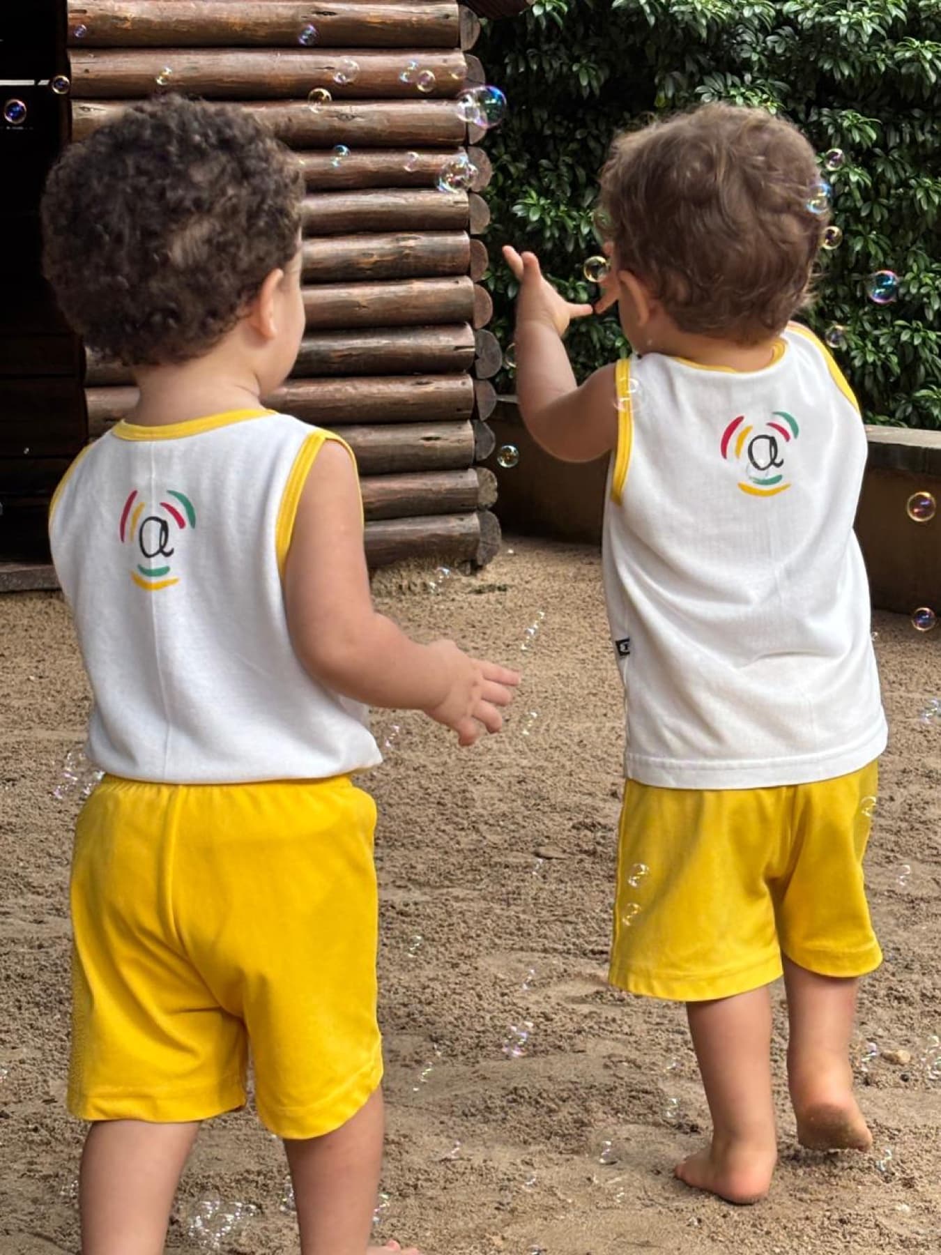 Duas crianças de uniforme Ateliê Aquarela brincando em frente à cabana de troncos no quintal